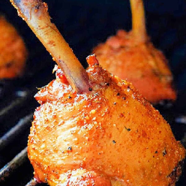 A close-up of a smoked chicken lollipop on a barbecue grill showcases its seasoned, crispy, golden-brown exterior. Two other drumsticks are visible in the background, slightly out of focus, adding depth to this mouthwatering scene.