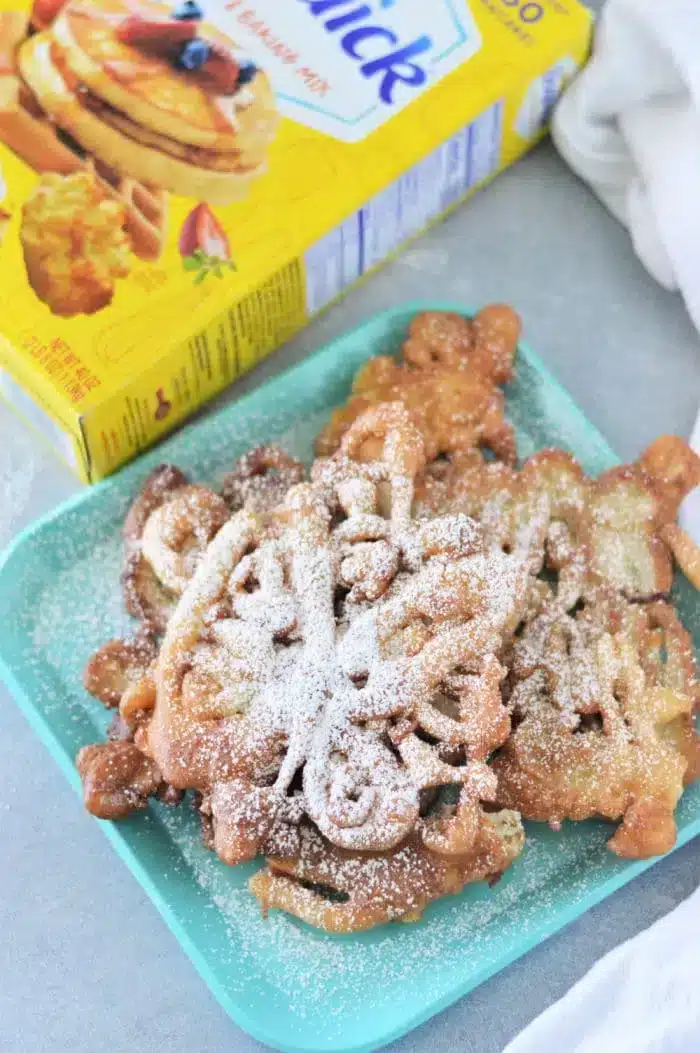 A plate with Bisquick funnel cakes dusted with powdered sugar sits enticingly next to a box of pancake mix.