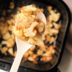 Close-up of roasted cauliflower florets on a spoon, showcasing the perfect transformation from frozen to crispy in an air fryer basket filled with more golden goodness.