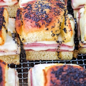 Close-up of several ham and cheese sliders on a cooling rack. Each slider is made with a soft roll, filled with sliced ham and melted cheese, and topped with poppy seeds. The golden-brown rolls look perfectly toasted, ideal for those who love crafting air fryer sandwiches.