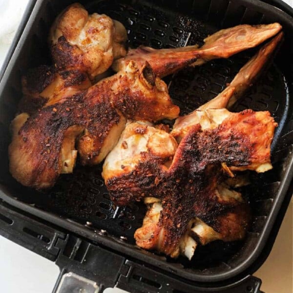 Air fryer turkey wings with crispy skin are cooked to perfection in an air fryer basket. The browned appearance of the wings indicates they are well-cooked. Placed on a white surface, a white cloth peeks out in the background, adding a touch of elegance to the scene.