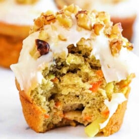 A partially eaten apple carrot muffin with frosting and chopped nuts rests on a white surface.