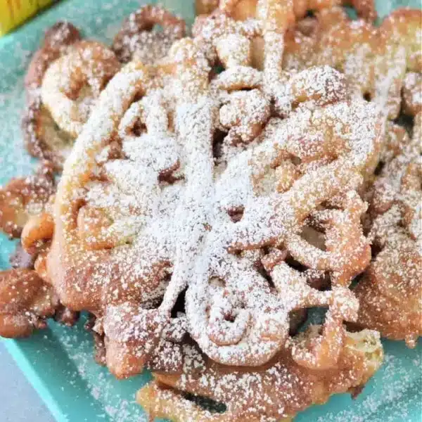 A plate of Bisquick funnel cakes dusted with powdered sugar rests on a teal square dish. A yellow box is partially visible in the background. The funnel cakes boast a light golden-brown color and intricate, swirling patterns.