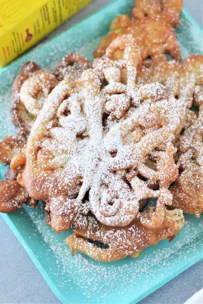 A plate of Bisquick funnel cakes dusted with powdered sugar rests on a teal square dish. A yellow box is partially visible in the background. The funnel cakes boast a light golden-brown color and intricate, swirling patterns.