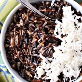 Bowl of black rice pudding topped with shredded coconut, placed on a plaid cloth, with a spoon.