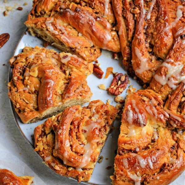 A spiral pecan cinnamon roll with icing, meticulously crafted like a bread wreath, features enticing slices on a white plate beside a bowl of pecans.