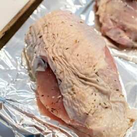 Raw turkey breast pieces seasoned with herbs rest on a foil-lined baking sheet, ready for roasting. Their marbled surface gleams with visible skin and spices. This method mirrors techniques seen in "How to Cook a Butterball Turkey Breast," ensuring tender, flavorful results each time.