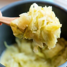 A wooden spoon holds a portion of buttered cabbage over a pot, cooked to softness in the pressure cooker. The cabbage appears lightly seasoned with a hint of butter, showcasing its pale green color. The pot is partially visible in the background.