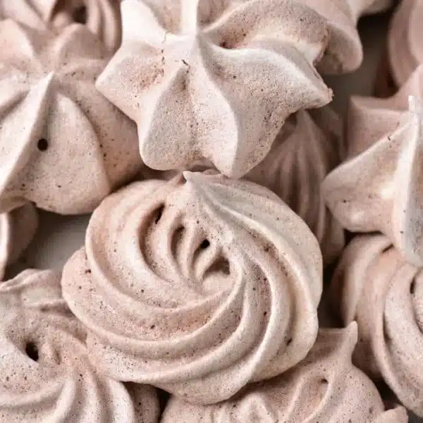 Close-up of several beige meringue cookies with a swirled and star-shaped design. These chocolate meringue cookies have a light, airy texture and are arranged in a pile, showcasing their intricate patterns and slight variations in shape and size.