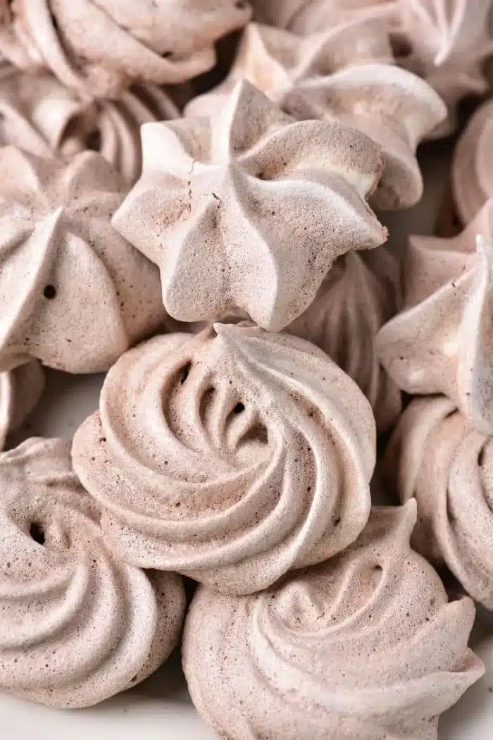 Close-up of several beige meringue cookies with a swirled and star-shaped design. These chocolate meringue cookies have a light, airy texture and are arranged in a pile, showcasing their intricate patterns and slight variations in shape and size.