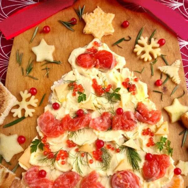 A festive Christmas butter board features a tree-shaped appetizer adorned with cheese, tomatoes, herbs, and crackers on a wooden board draped with a red and white cloth.