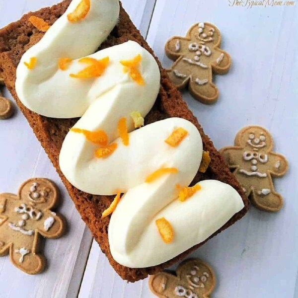 Copycat Starbucks gingerbread loaf topped with whipped cream and orange zest, surrounded by gingerbread cookies on a white background.