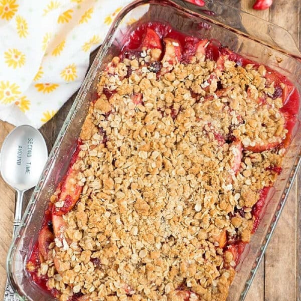 A glass dish of cranberry apple cobbler with oat topping, surrounded by fruit and a yellow-patterned towel.