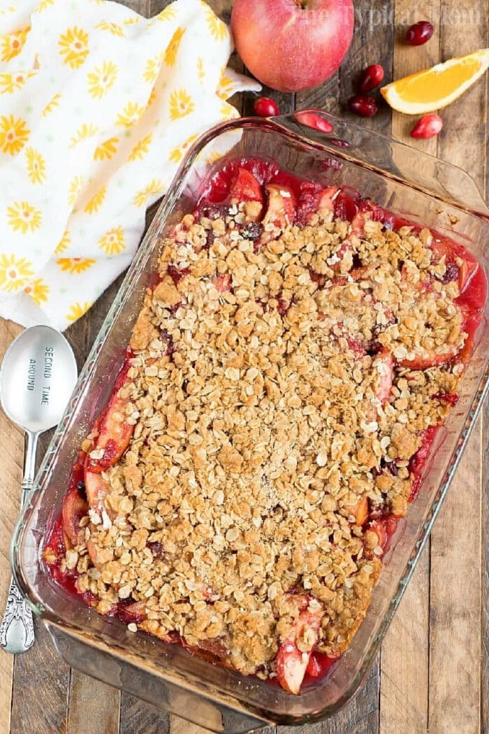 A glass dish of cranberry apple cobbler with oat topping, surrounded by fruit and a yellow-patterned towel.