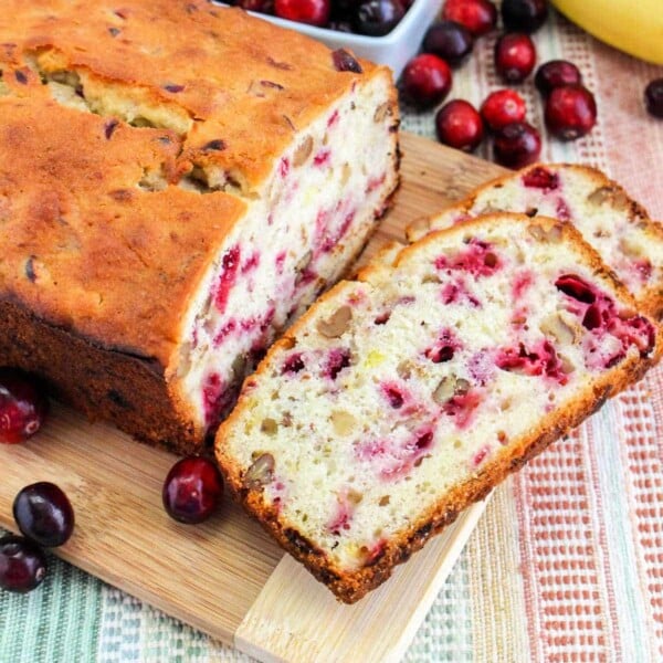 Banana cranberry bread sits invitingly on a cutting board, flanked by vibrant slices surrounded by fresh cranberries and a ripe banana.