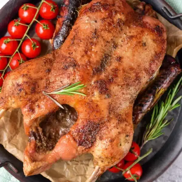 Roasted whole duck, beginning its journey as a frozen delight, sits on a tray with herbs and charred sausages. Cherry tomatoes on the vine garnish the side. The tray rests elegantly on a green cloth against a neutral backdrop.