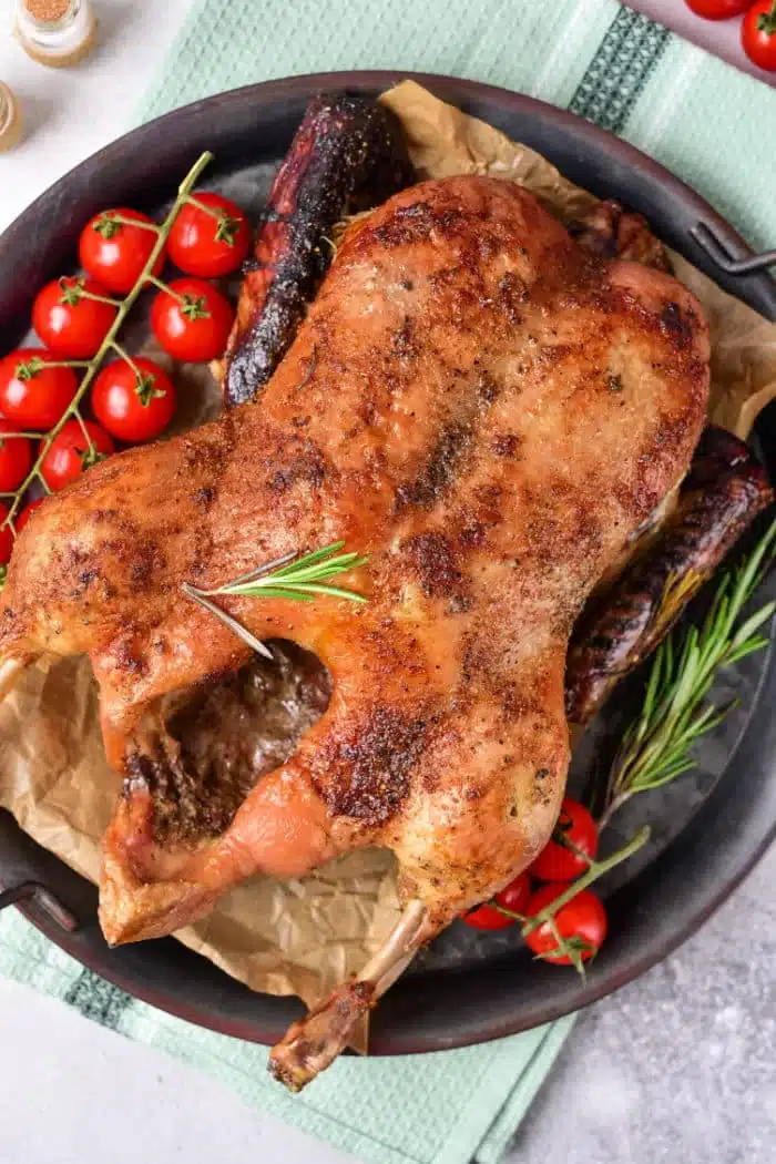 Roasted whole duck, beginning its journey as a frozen delight, sits on a tray with herbs and charred sausages. Cherry tomatoes on the vine garnish the side. The tray rests elegantly on a green cloth against a neutral backdrop.