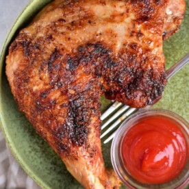 A cooked chicken leg is served on a green plate with a small glass bowl of ketchup, offering inspiration for those wondering how to cook turkey legs and thighs. A fork rests beneath the succulent meat, and a small bowl with a white substance peeks from the bottom left corner.