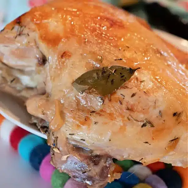 A piece of roasted turkey breast with a golden brown, crispy skin and herbs, including a bay leaf, rests on a colorful wool ball trivet. The scene is reminiscent of learning how to cook a Butterball turkey breast. A multicolored fabric subtly graces the background.