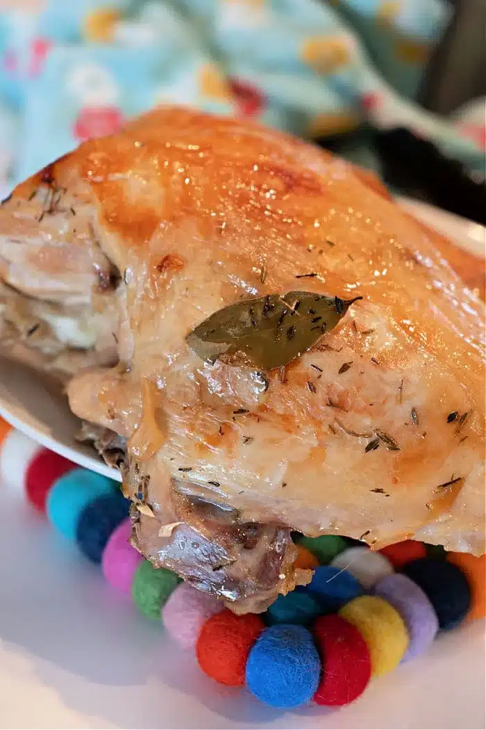 A piece of roasted chicken with a golden brown, crispy skin and herbs, including a bay leaf, rests on a colorful wool ball trivet. The scene is reminiscent of learning how to cook a Butterball turkey breast. A multicolored fabric subtly graces the background.