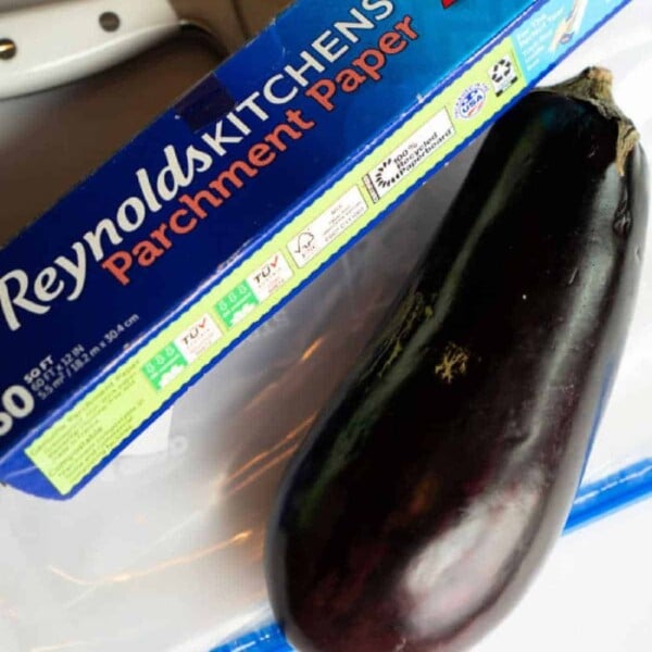 An eggplant sits beside a box of Reynolds Kitchens parchment paper on a pristine white surface, ready for your next culinary adventure. Discover how to freeze eggplant effectively using this kitchen essential.