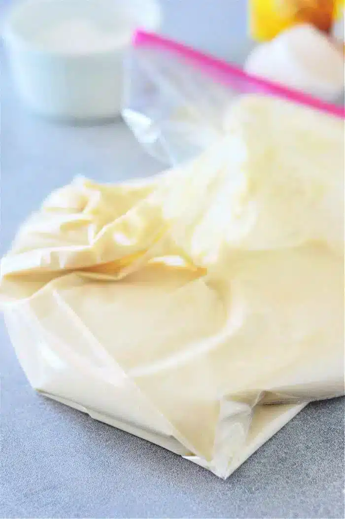 A clear plastic bag filled with light yellow Bisquick dough sits on a gray surface, ready for funnel cake magic. The bag appears sealed, and there are blurred objects in the background, possibly kitchen items.