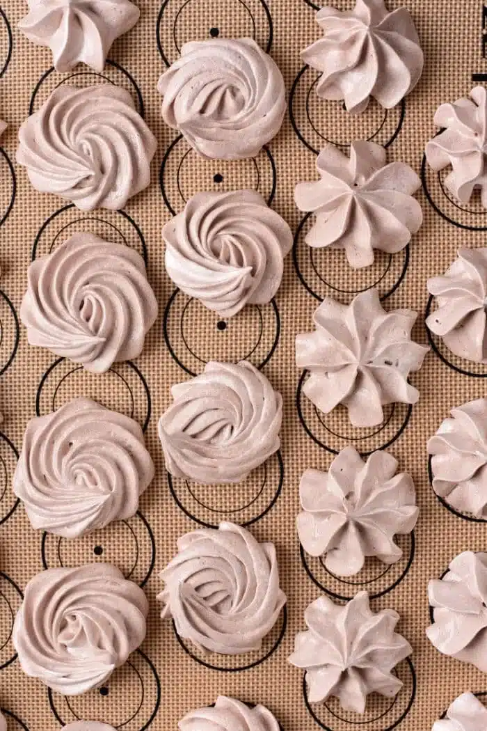 Rows of light brown chocolate meringue cookies are arranged on a baking mat with circular guides. The cookies are shaped in swirls and star patterns, boasting a smooth texture and slightly glossy appearance.