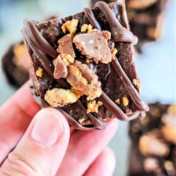 Close-up of a hand holding a decadent chocolate fudge, drizzled with rich chocolate and topped with crushed candy. The fudgy texture, enhanced by hints of peanut butter and creamy condensed milk, makes it a delightful indulgence.