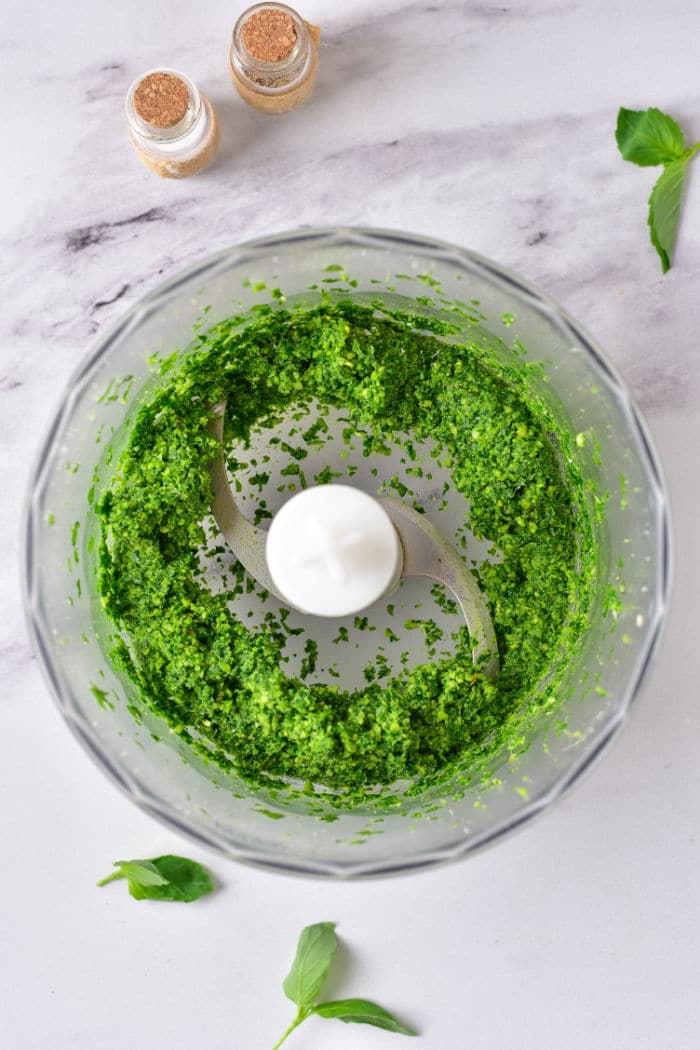 Green pesto without pine nuts swirls in a food processor bowl on a marble countertop, accompanied by fresh basil leaves and spice jars nearby.