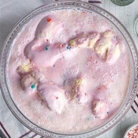 A bowl of pink ice cream punch, inspired by the ultimate Pink Punch Recipe, is adorned with colorful sprinkles on a wooden table. Beside it lies a striped napkin and silver spoons, ready for indulgence.