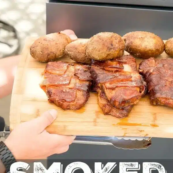 A person holds a wooden board displaying three bacon-wrapped smoked pork chops and four baked potatoes in front of a smoker grill. The text at the bottom reads Smoked Bacon Wrapped Chops.