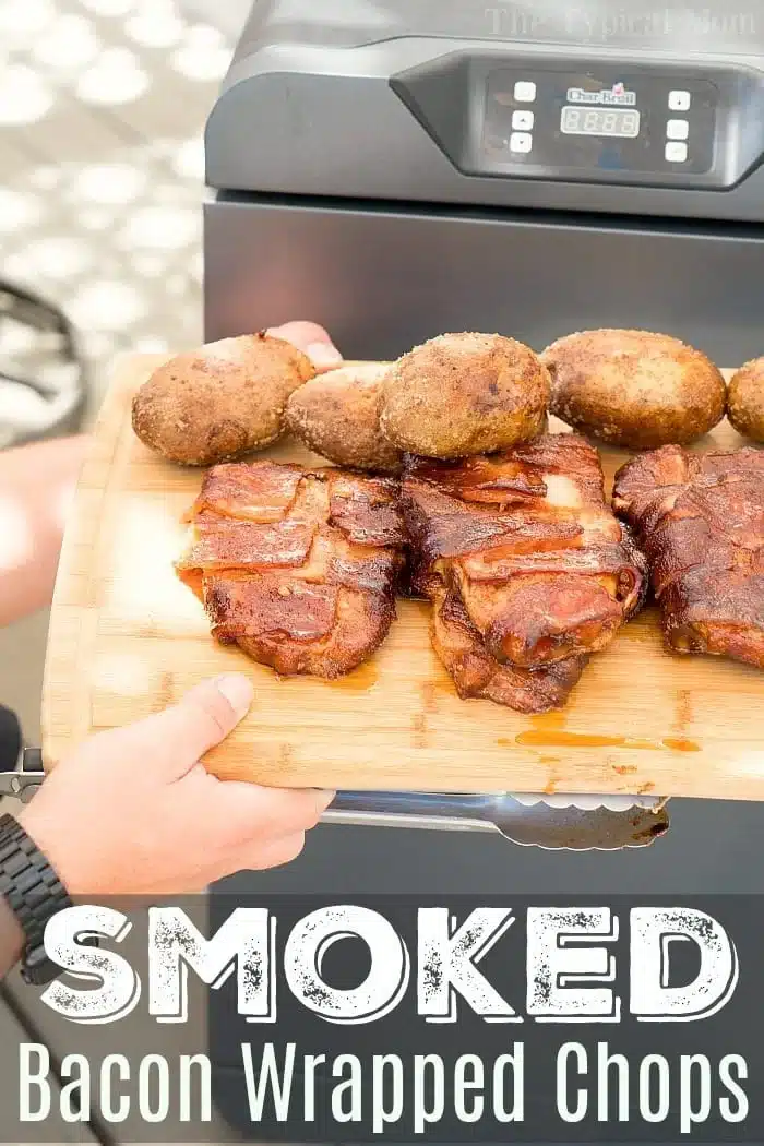 A person holds a wooden board displaying three bacon-wrapped smoked pork chops and four baked potatoes in front of a smoker grill. The text at the bottom reads Smoked Bacon Wrapped Chops.