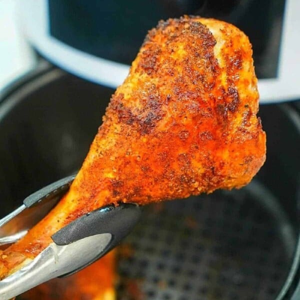 A cooked turkey leg with a seasoned, crispy coating is held by tongs over an air fryer basket, with another turkey leg visible inside the air fryer.