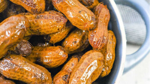 A bowl of Crockpot boiled peanuts sits on a wooden surface, complemented by a gray cloth in the background.