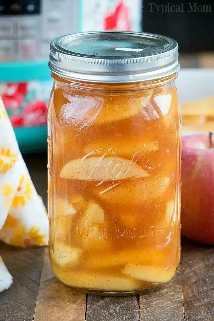 A glass jar filled with slices of apple in syrup sits on a wooden surface, evoking the sweetness of a Bisquick funnel cake. An apple and a floral-patterned cloth are nearby, completing the cozy scene.