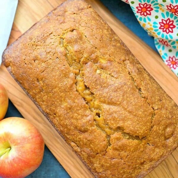 A loaf of easy pumpkin apple bread rests on a wooden board, surrounded by fresh apples and a colorful towel.