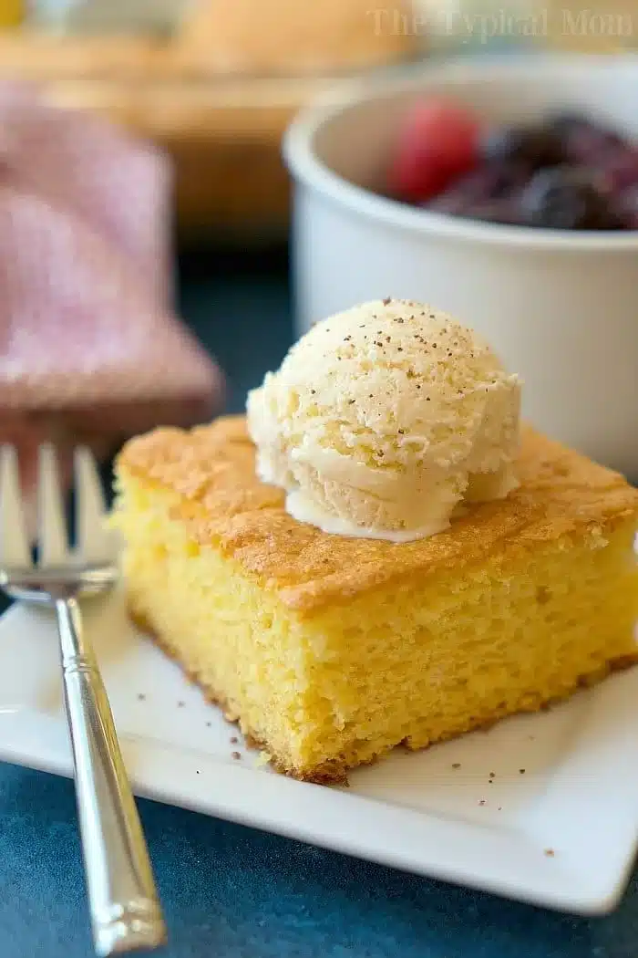 A slice of cornbread topped with a scoop of vanilla ice cream sits elegantly on a white plate with a fork, reminiscent of an egg nog pudding cake in its rich allure. A bowl of fruit is artistically blurred in the background.