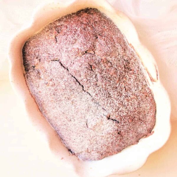 A freshly baked loaf of chocolate gingerbread bread in a scalloped baking dish, dusted with flour.