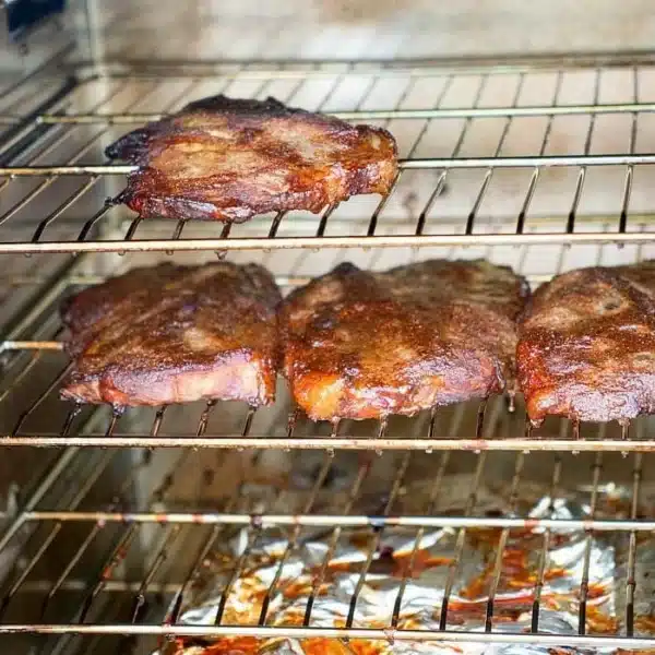 Four pieces of smoked pork steaks with dry rub are placed on two smoker racks, with a foil-lined tray catching drippings below. The meat appears perfectly browned, indicating it has been roasted or broiled to smoky perfection.