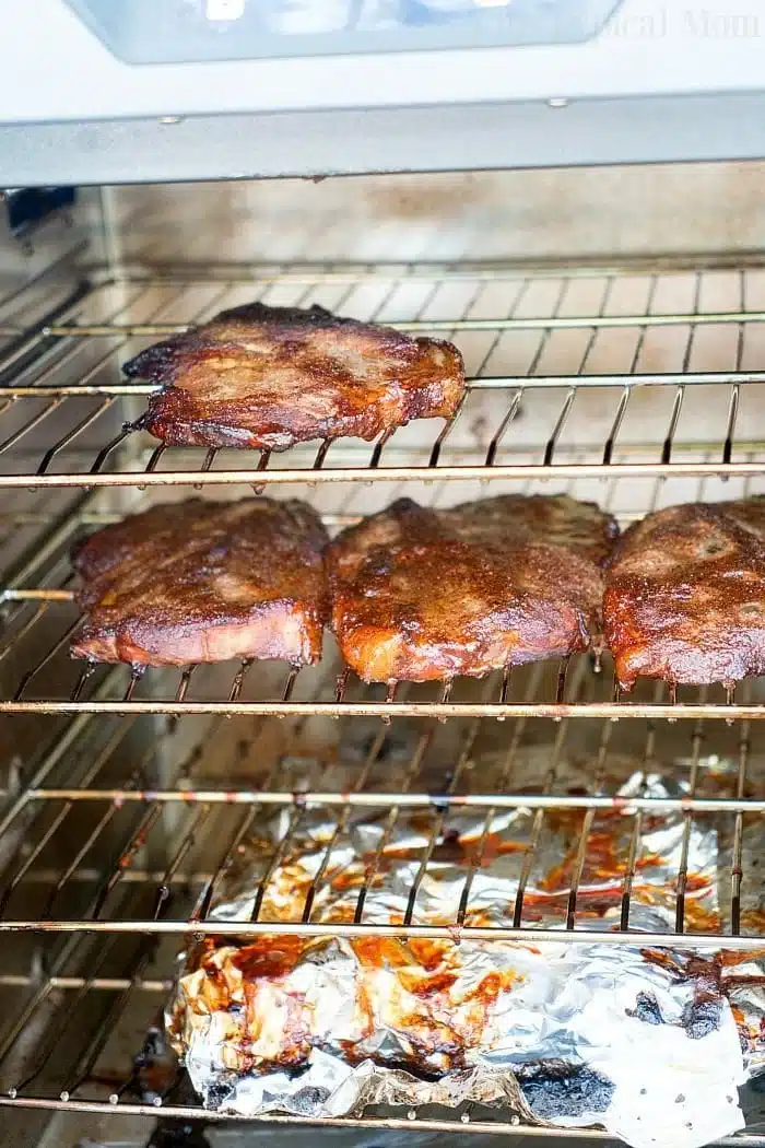 Four pieces of smoked pork steaks with dry rub are placed on two smoker racks, with a foil-lined tray catching drippings below. The meat appears perfectly browned, indicating it has been roasted or broiled to smoky perfection.