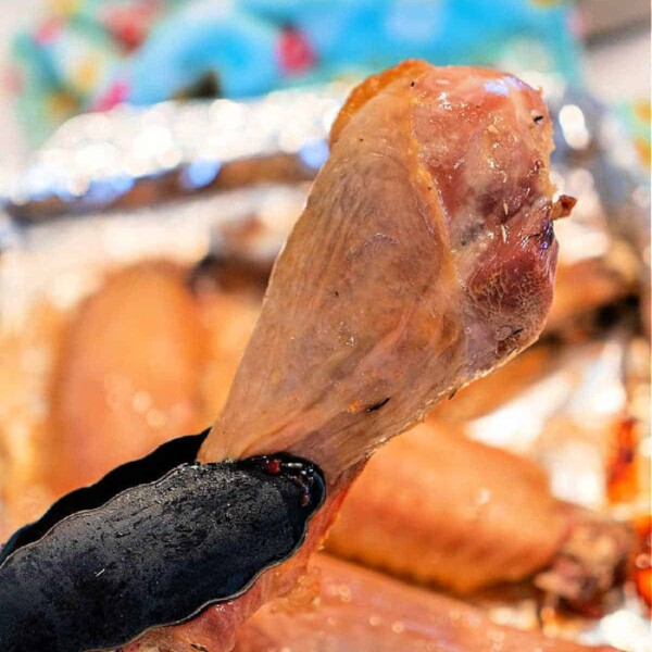 Close-up of a baked turkey leg being held by black tongs. The turkey leg is golden brown with a slight sheen, and several more baked turkey legs are visible in the blurred background on a foil-covered tray. A colorful cloth is partially visible.