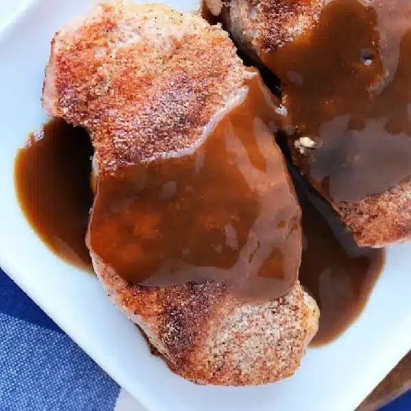 Two pieces of seasoned pork chops, expertly cooked from frozen, sit on a white plate, topped with a generous amount of brown gravy. The dish rests on a blue and white checkered tablecloth, ready to delight the senses.