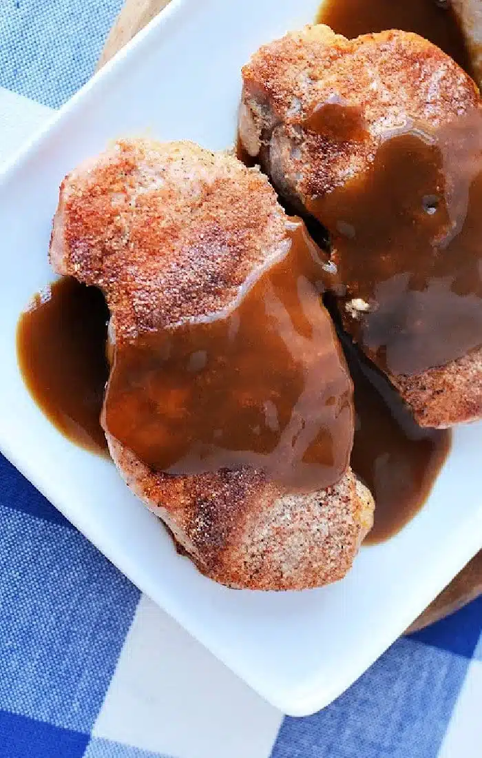 Two cooked pork chops with gravy sit on a white plate, the result of expertly cooking frozen pork chops. The plate rests on a surface adorned with a blue and white checked pattern.