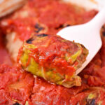 An Instant Pot stuffed cabbage roll, covered in tomato sauce, is lifted by a white spoon from a dish of similar rolls.