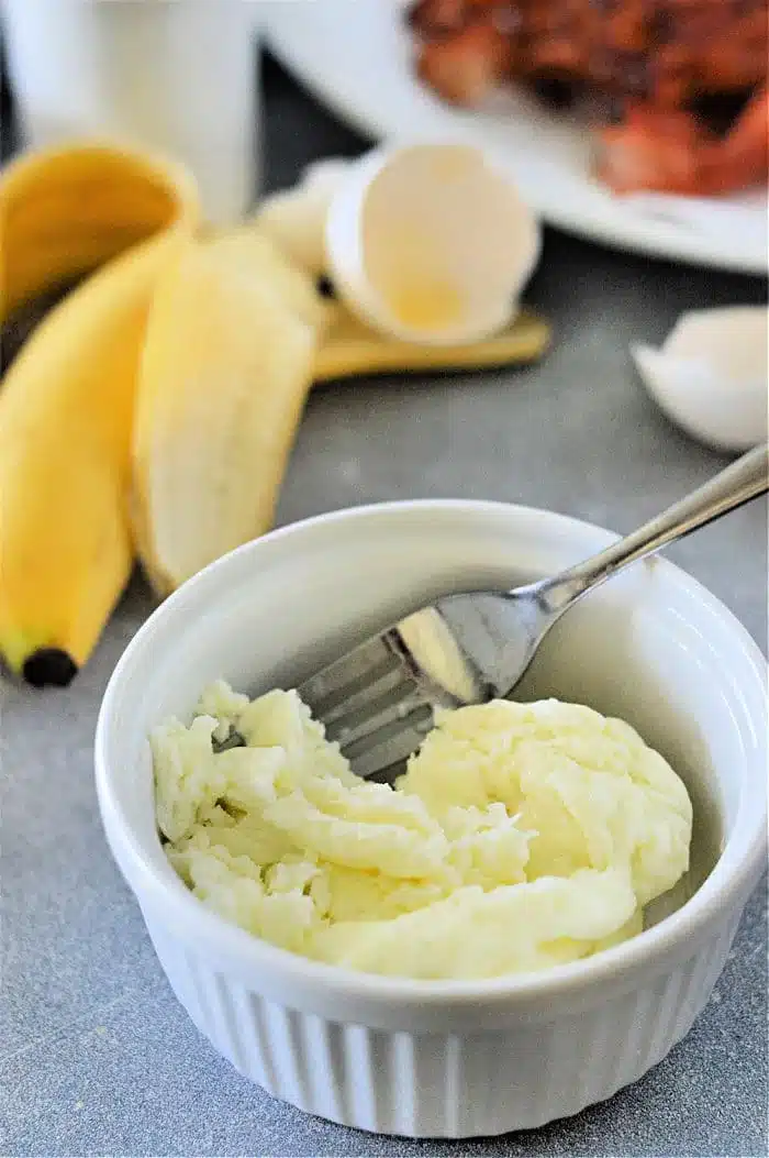 A small white bowl contains cooked egg whites with a fork resting inside. In the background, there are two unpeeled bananas, an empty eggshell from cooked egg whites, and a plate of crispy bacon on a gray surface.