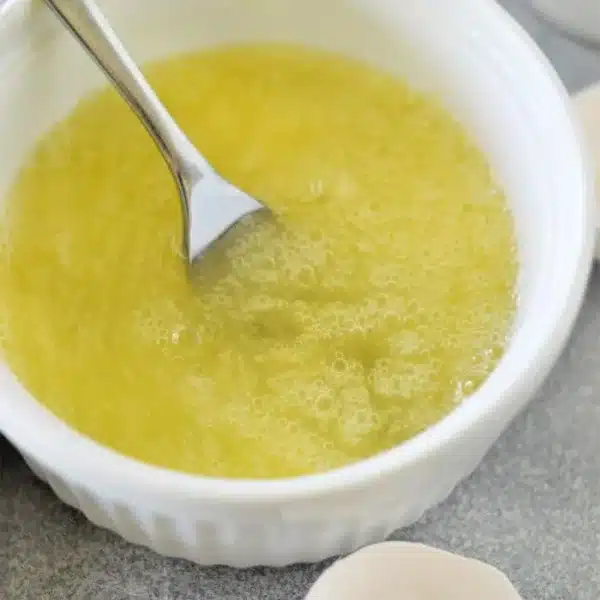 A bowl containing beaten egg whites with a fork lies on a gray countertop. Two empty eggshell halves are nearby.