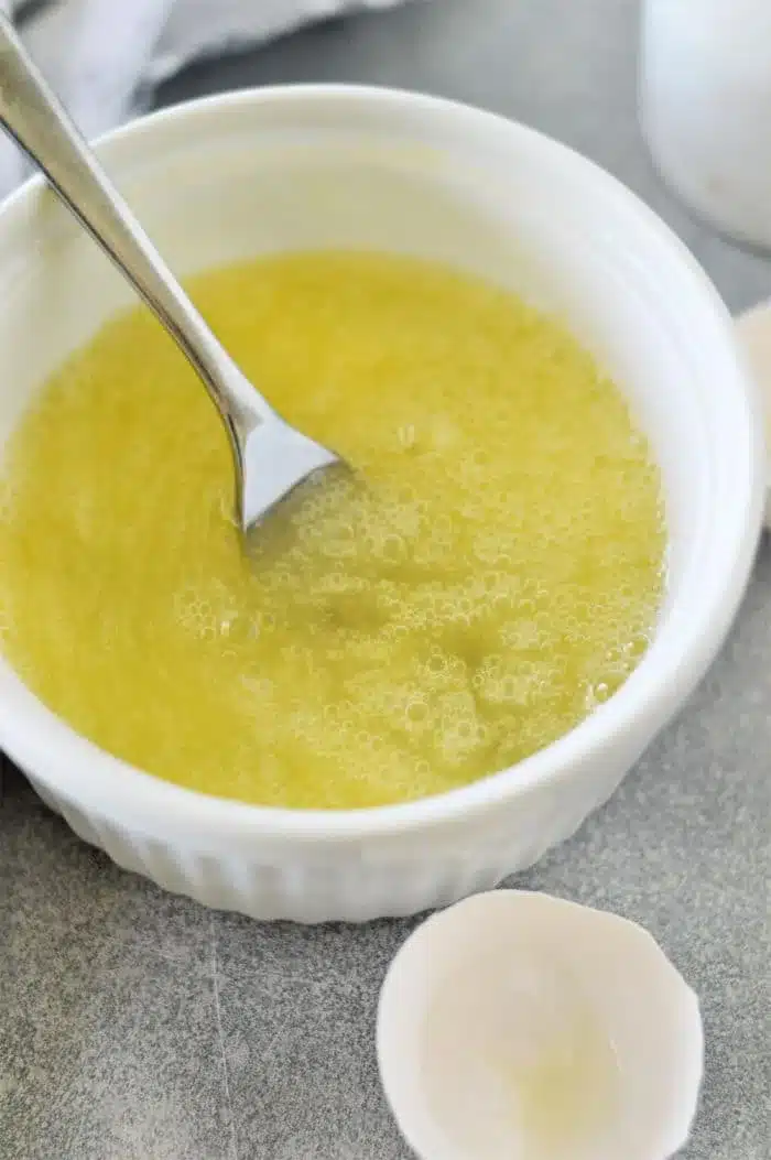 A white ceramic bowl containing beaten eggs with a fork resting inside sits ready for the next step in how to cook egg whites. Eggshell halves lie scattered nearby on the gray surface, hinting at the simple elegance of this culinary process.