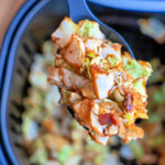 Close-up of a spoon holding savory fried cabbage with crispy bacon pieces, over an air fryer basket.