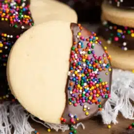 A close-up of a homemade shortbread cookie half-dipped in chocolate and coated with colorful sprinkles. The cookie rests on a textured white surface, surrounded by more cookies and chocolate chips in the background.