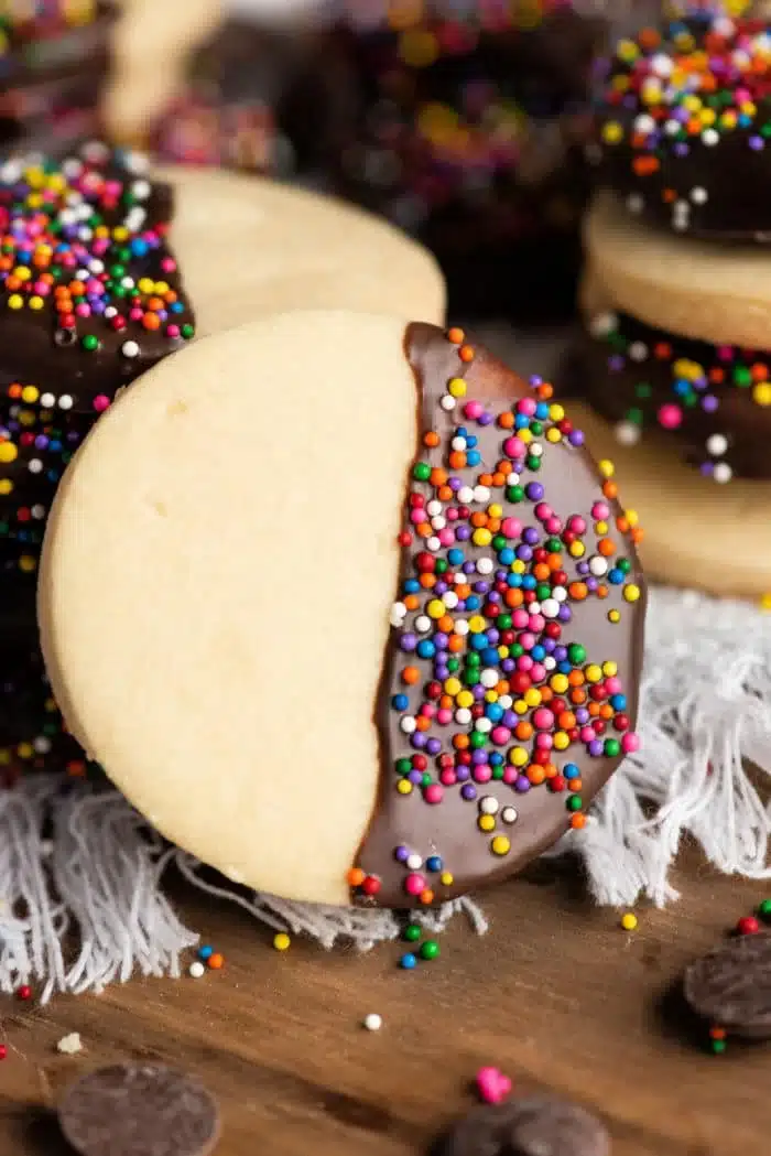 A close-up of a homemade shortbread cookie half-dipped in chocolate and coated with colorful sprinkles. The cookie rests on a textured white surface, surrounded by more cookies and chocolate chips in the background.